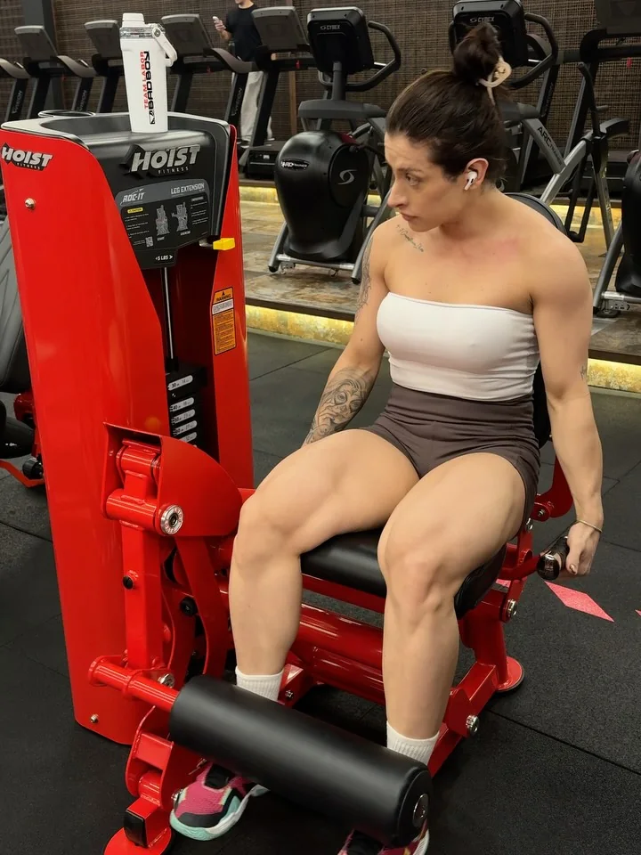Laura Carnecine sentada na cadeira extensora vermelha da academia, de top branco e shorts marrom, olhando para o lado