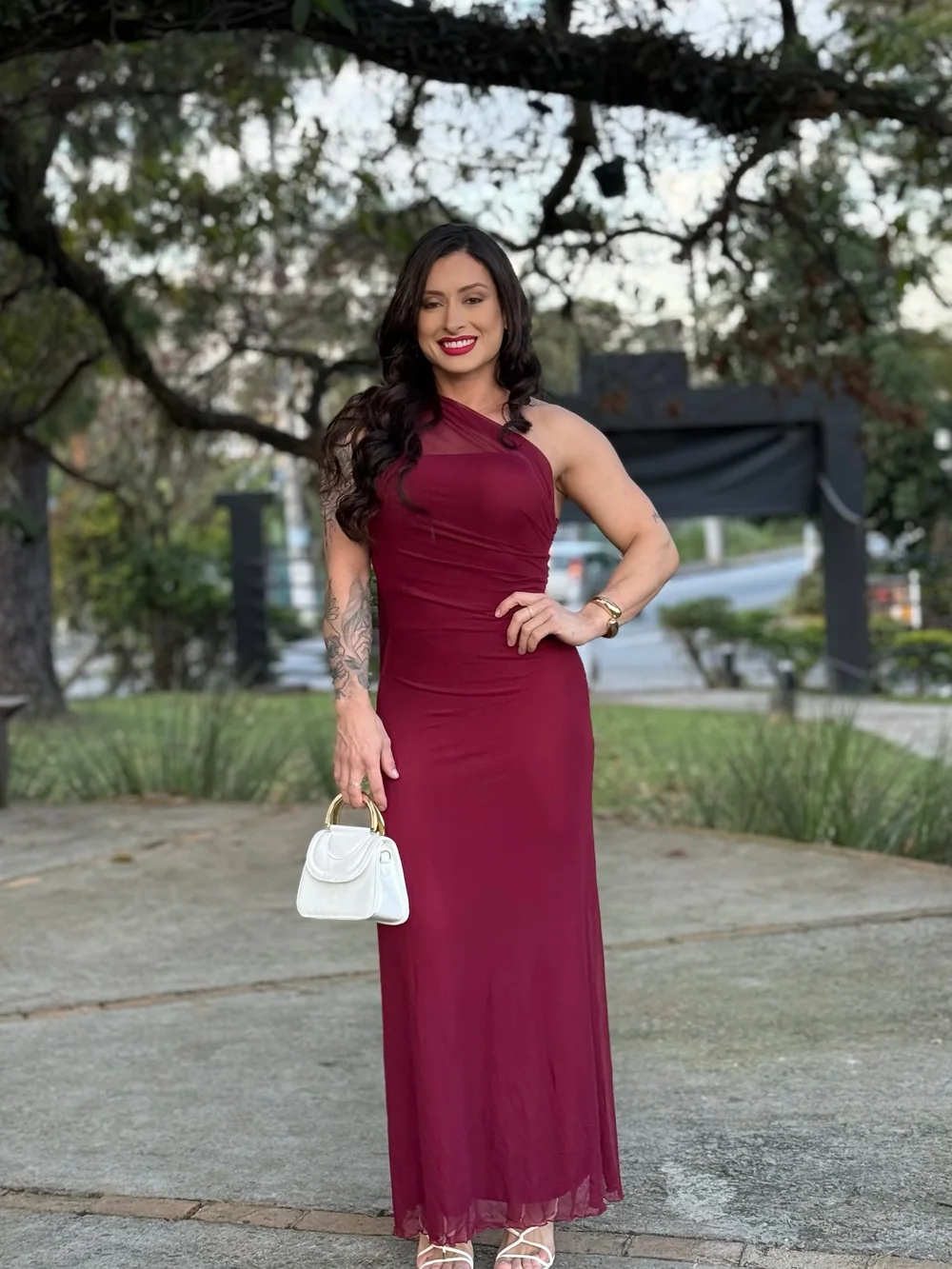Laura Carnecine de vestido longo bordô e bolsa branca, com a mão na cintura, ao ar livre sob uma árvore